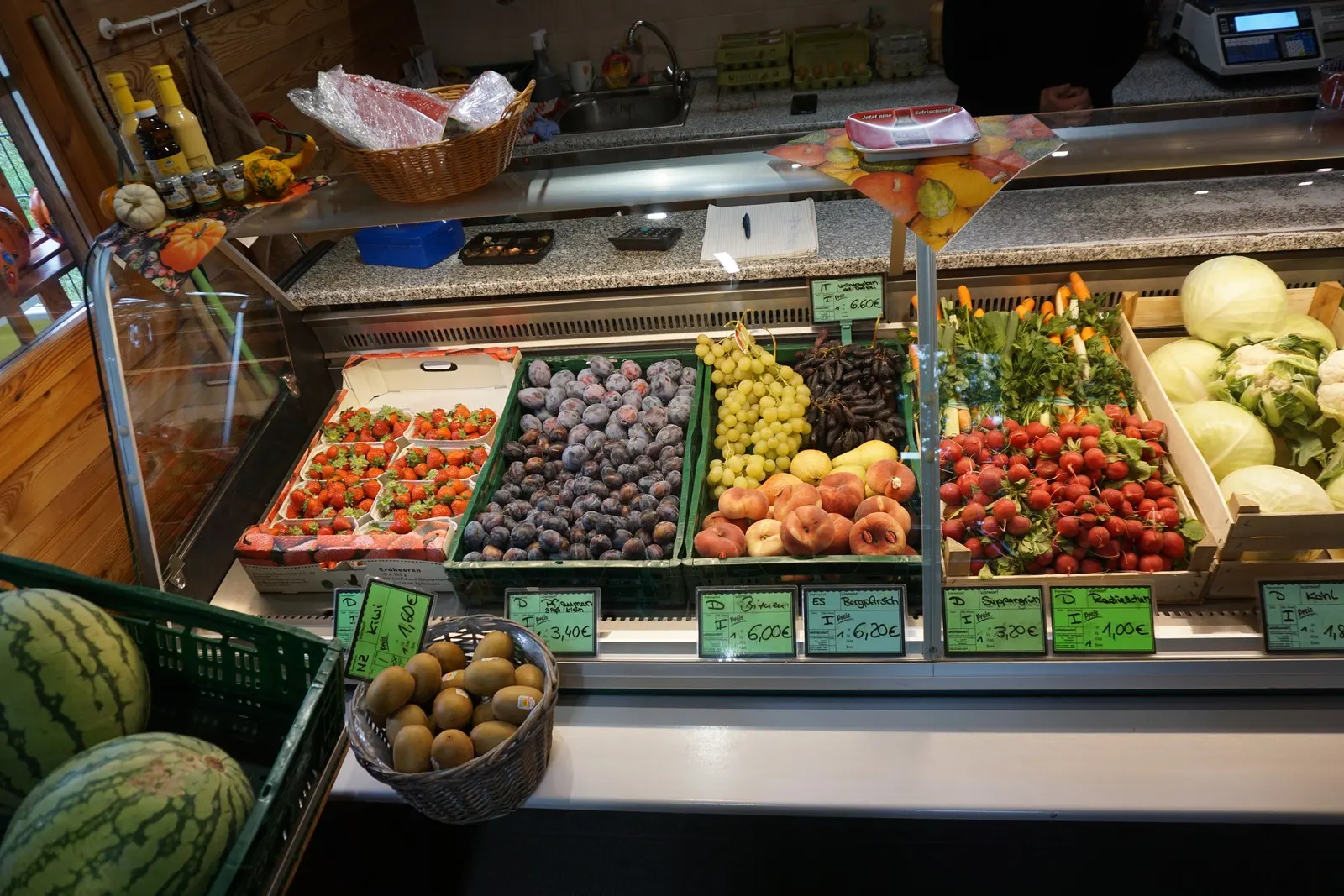 Obst und Gemüse in der Theke, davor Melonen und Kiwis